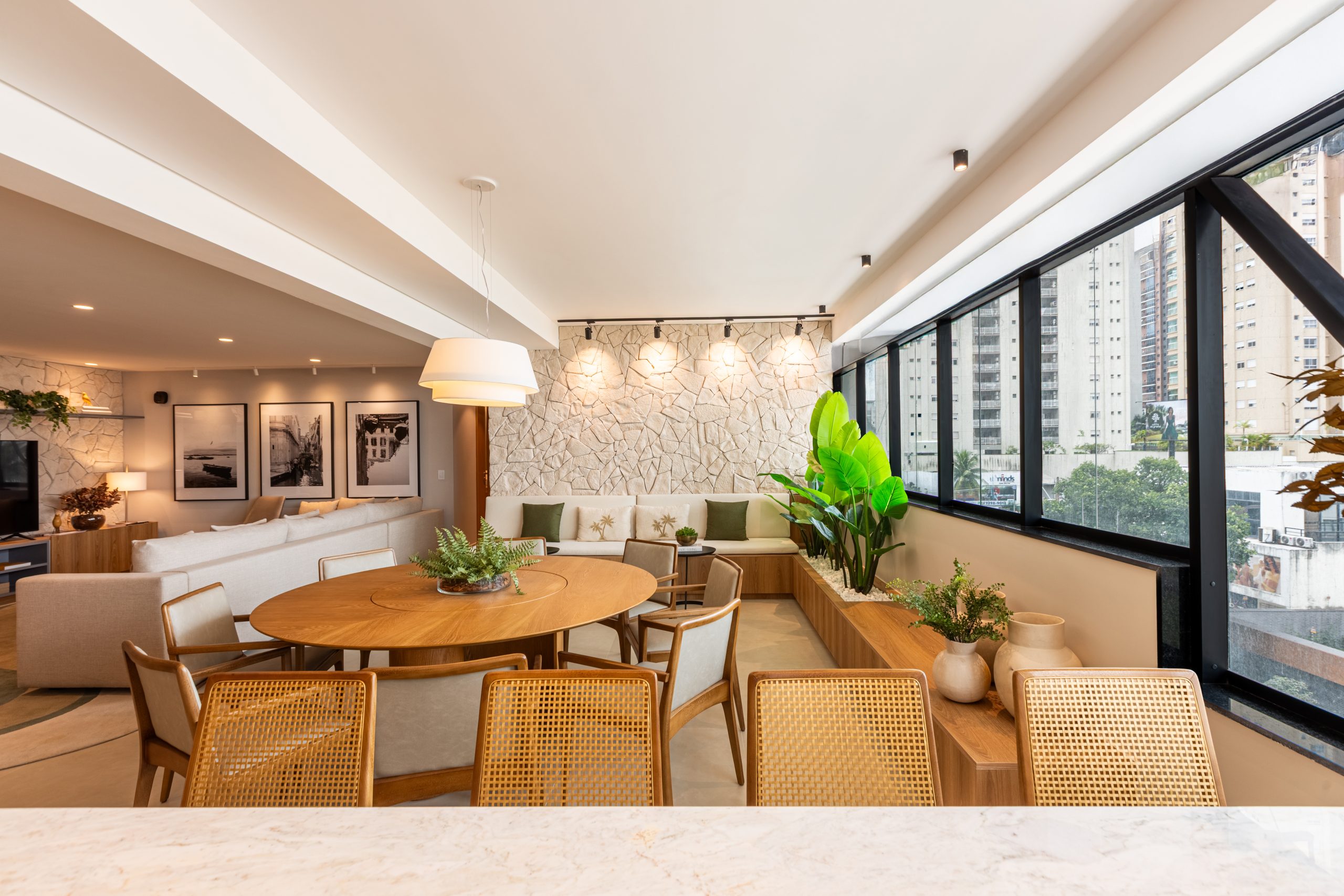 Fechamento de Varanda Gourmet - sala de estar integrada com varanda e cozinha - aconchegante com pegada de design natural, utilização de pedra na parede, marcenaria e mobiliário personalizado com revestimento de madeira nas paredes, decoração em tons neutros - mesa de jantar redonda para 8 lugares