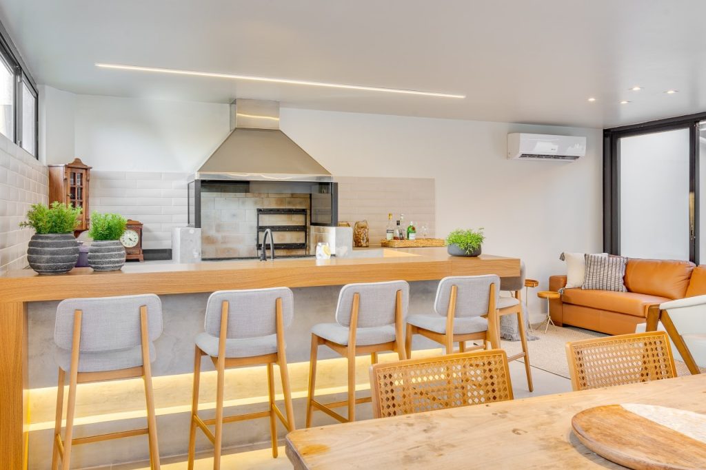 Espaço gourmet moderno e integrado com grande bancada iluminada por LED com banquetas cinzas. Ao fundo, a área da churrasqueira conta com revestimento de tijolinhos brancos e uma coifa de aço inoxidável em destaque.