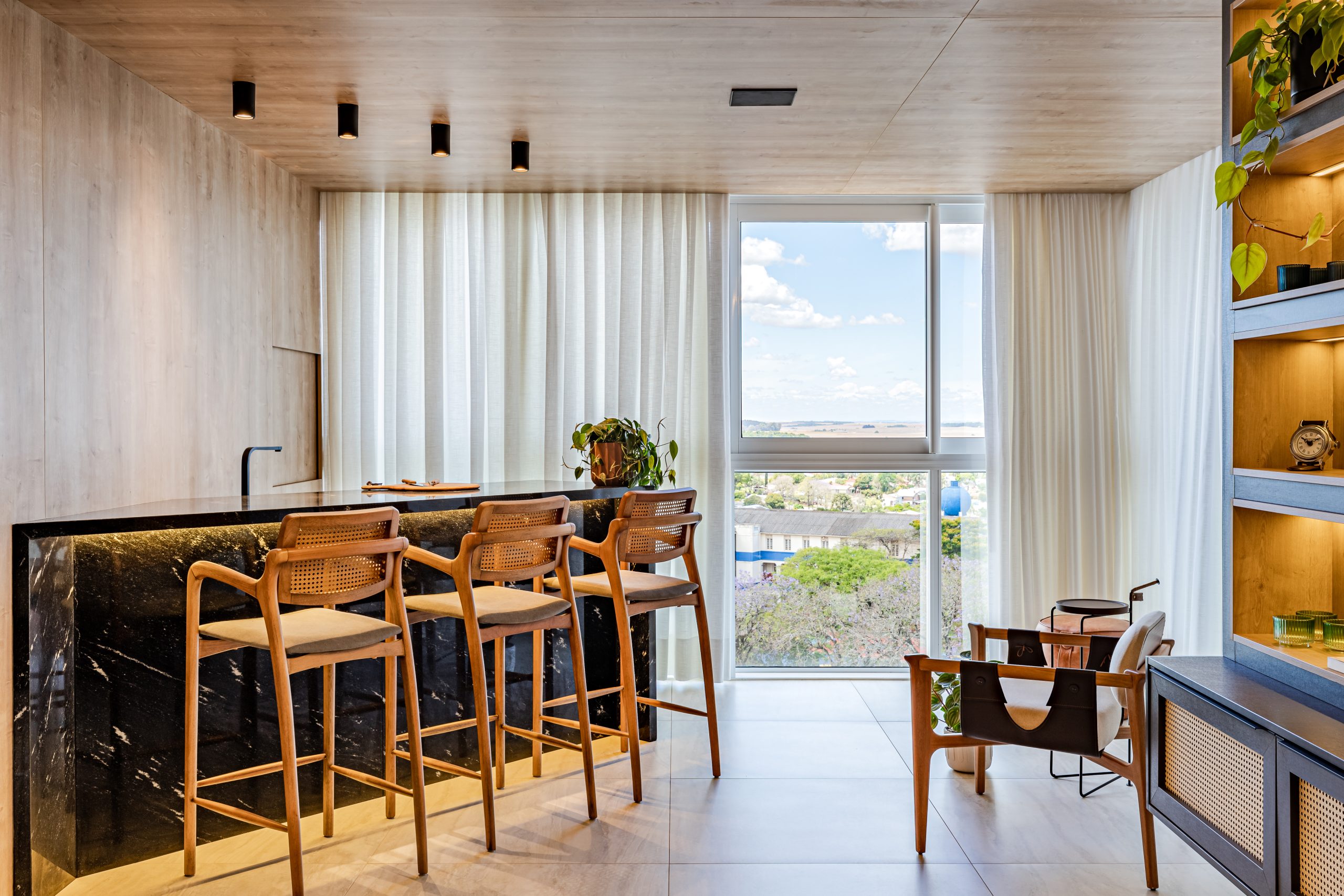 Espaço gourmet de apartamento com bancada em mármore escuro com iluminação inferior e banquetas de madeira com palhinha