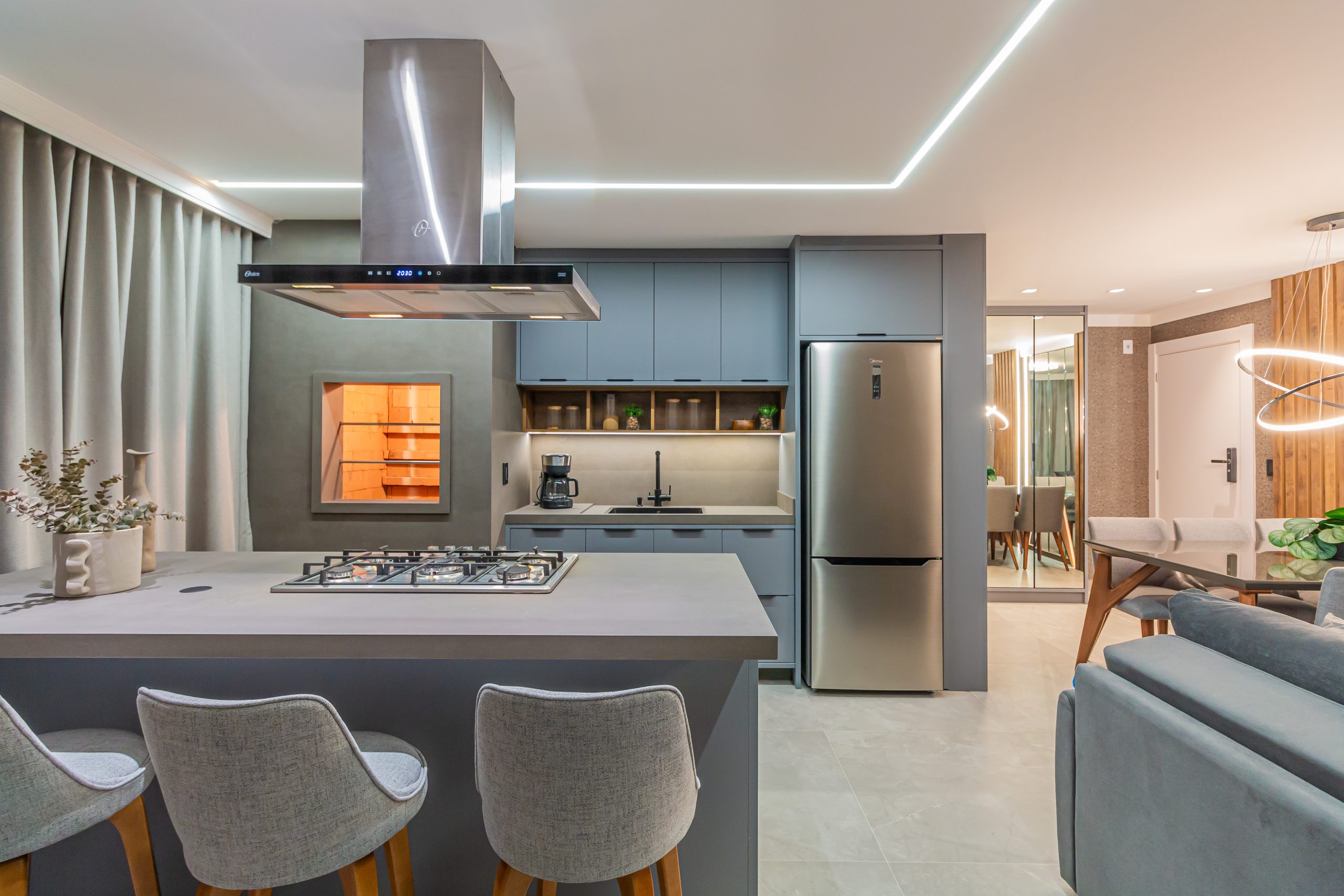 Espaço gourmet moderno com paleta de tons cinza nos armários. Ilha central, valorizada pela iluminação em perfil de LED no teto. A ilha integra o cooktop a uma coifa de inox de design marcante, servindo também como bancada de apoio com confortáveis banquetas estofadas. Ao fundo, a churrasqueira embutida e os eletrodomésticos em aço escovado garantem funcionalidade e sofisticação à área de convivência. Cozinha moderna