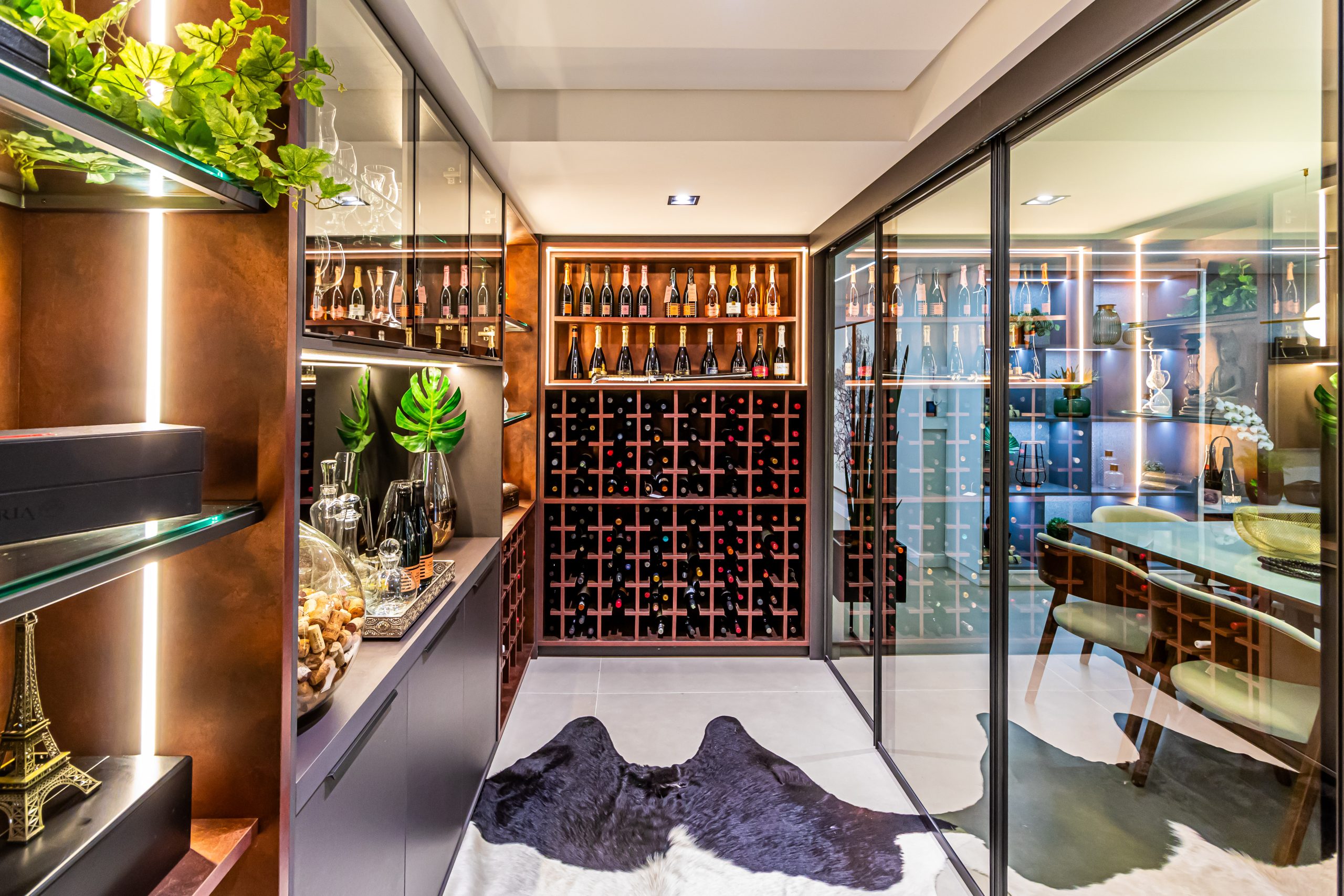 adega integrada com sala de estar. Foto de uma adega de vinhos ampla, de luxo com móveis planejados em cores escuras com detalhe em acabamento de aço corten. A foto mostra áreas onde as garrafas de vinho estão expostas deitadas mas também áreas com portas de vidro translúcidas para ver copos e taças. A adega fica separada por portas de vidro que dividem o ambiente com a sala de jantar.