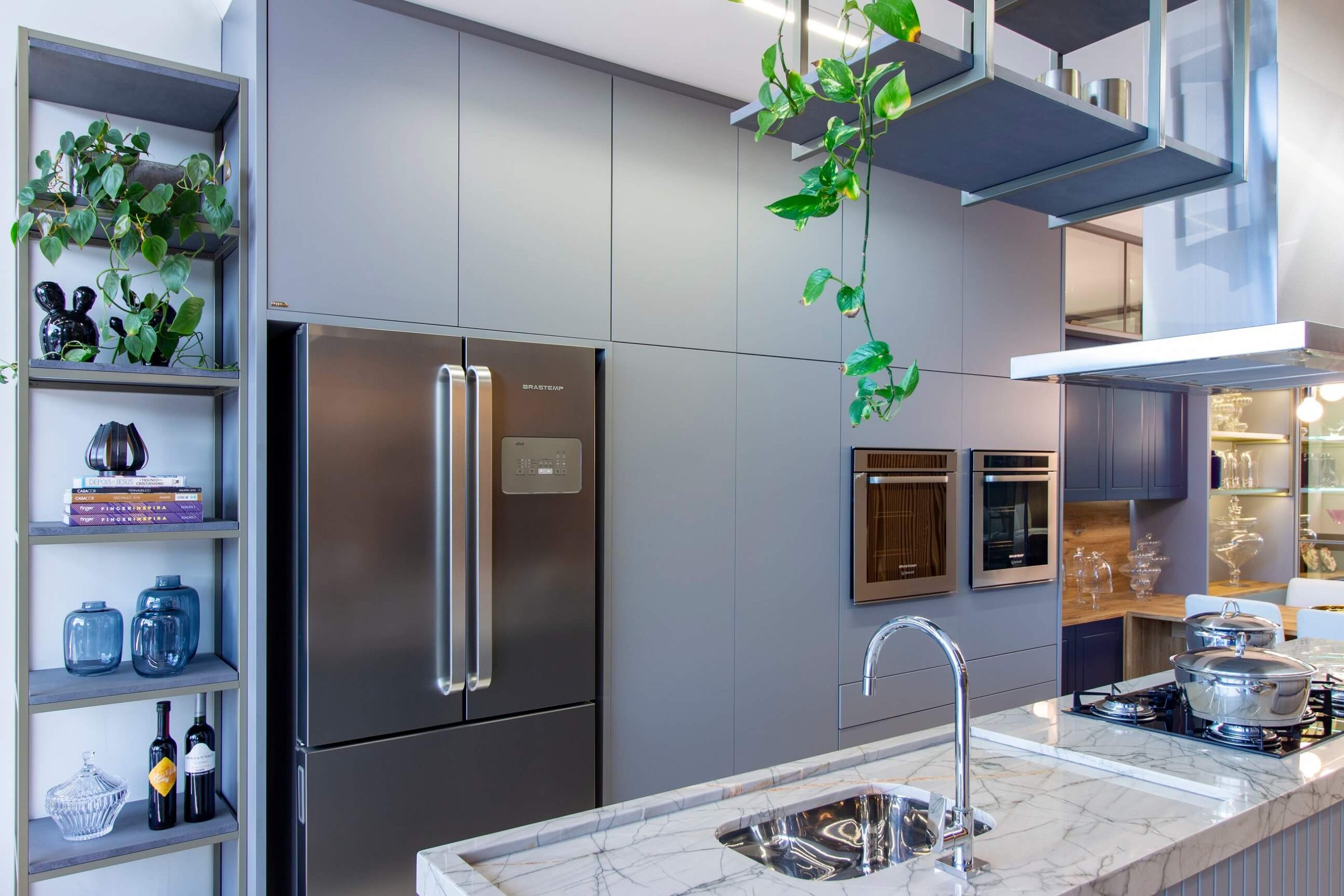 Cozinha Contemporânea com Ilha em tom de cinza e azul escuro. As portas são lisas e a cozinha possui torre quente onde forno e microondas ficam lado a lado. Sobre a área da pia, há uma estrutura metálica com prateleiras que estão decoradas com plantas e objetos minimalistas.