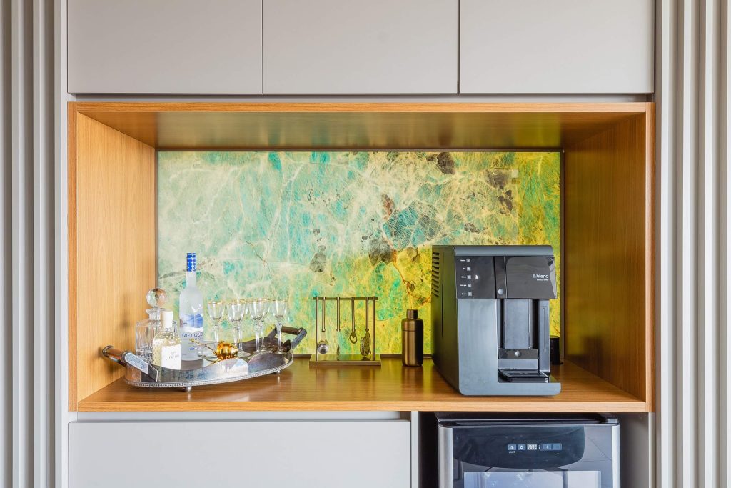 Detalhe de um nicho para cozinha, com cantinho para o café e espaço de mini bar com adega. A composição é em tons neutros com uma moldura amadeirada por dentro do nicho e um fundo com marmore iluminado dá o charme a decoração. 