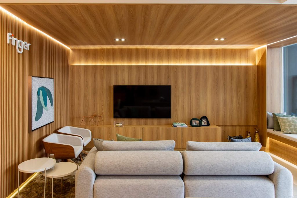 Sala de Estar ampla em ambiente personalizado, com móveis planejados em tom de madeira mel que revestem o painel de TV e as paredes, seguindo também o teto, com iluminação em LED de forma indireta. O sofá é claro e a decoração é leve. Na lateral tem duas poltronas individuais e mesas laterais na cor branca.