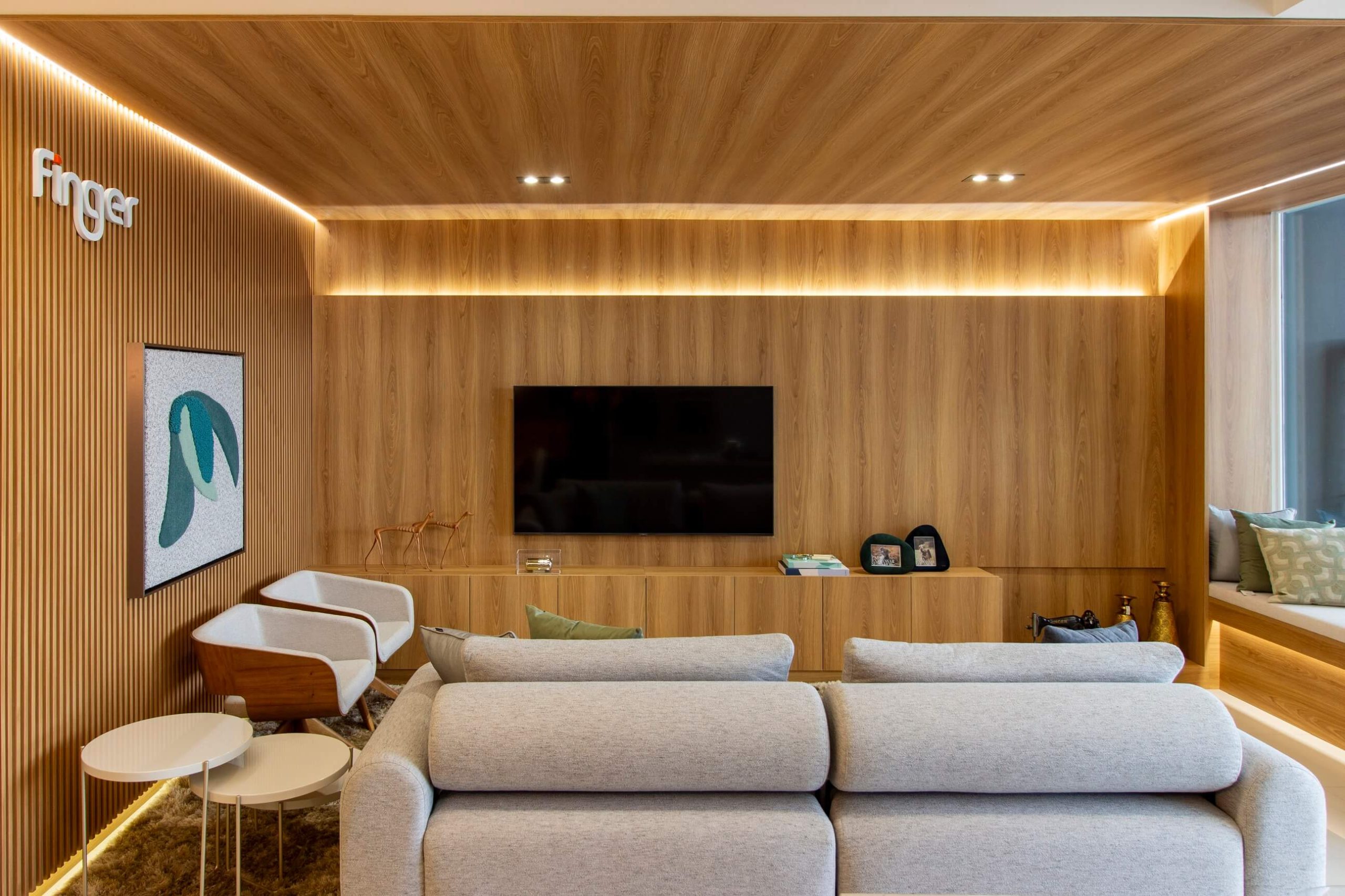 Sala de Estar ampla em ambiente personalizado, com móveis planejados em tom de madeira mel que revestem o painel de TV e as paredes, seguindo também o teto, com iluminação em LED de forma indireta. O sofá é claro e a decoração é leve. Na lateral tem duas poltronas individuais e mesas laterais na cor branca.