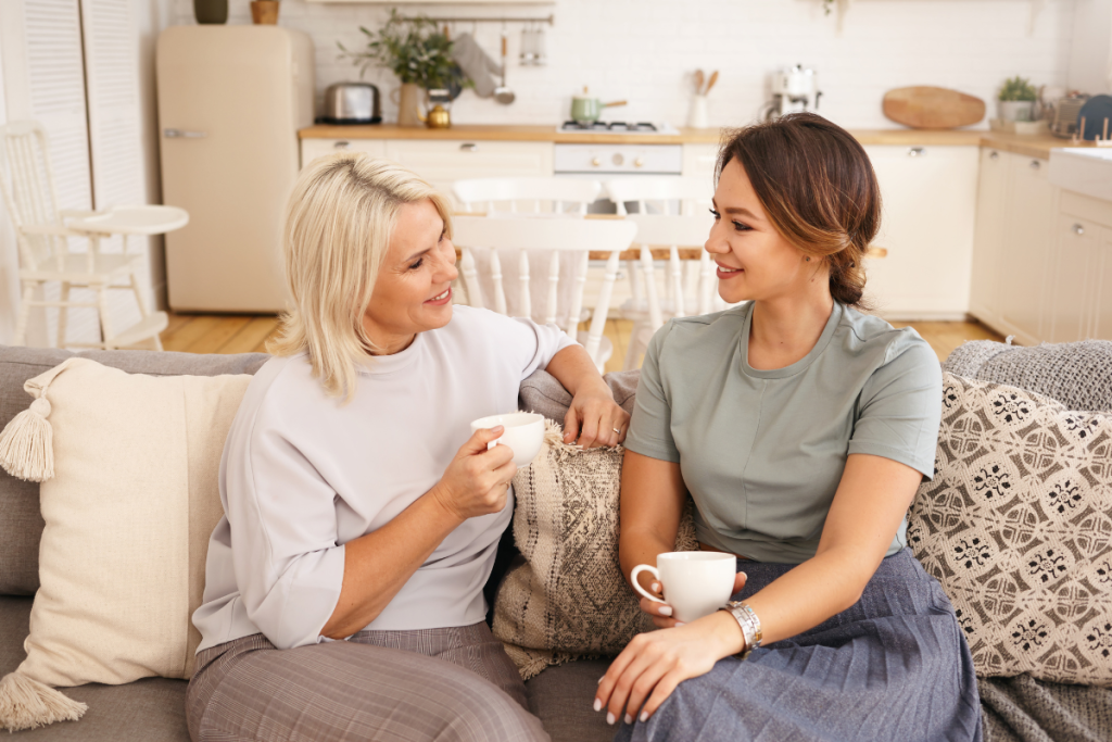 tendencia-casa-de-vo-aconchego-e-nostalgia-presentes-na-decoracao-afetiva-1. A foto mostra duas mulheres de gerações diferentes conversando e tomando chá, sentadas no sofá. Ao fundo vê-se a cozinha planejada em tons claros com detalhe em madeira.