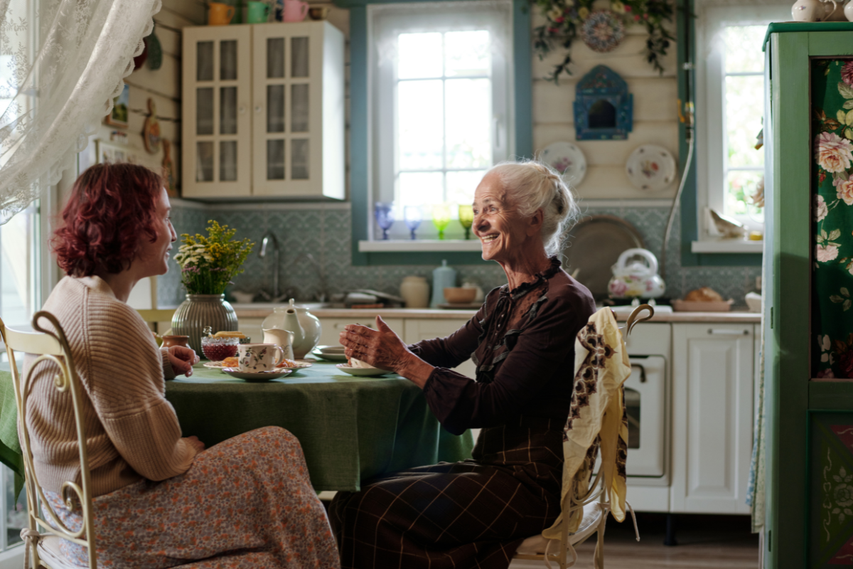 tendência casa de vó. Imagem mostra duas mulheres conversando sentadas em uma cozinha, tomando um chá. O semblante das mulheres é feliz. Uma delas é uma senhora de mais idade e a outra uma mulher em torno de 50 anos, aparentemente mãe e filha curtem a casa e o momento de confraternização. 