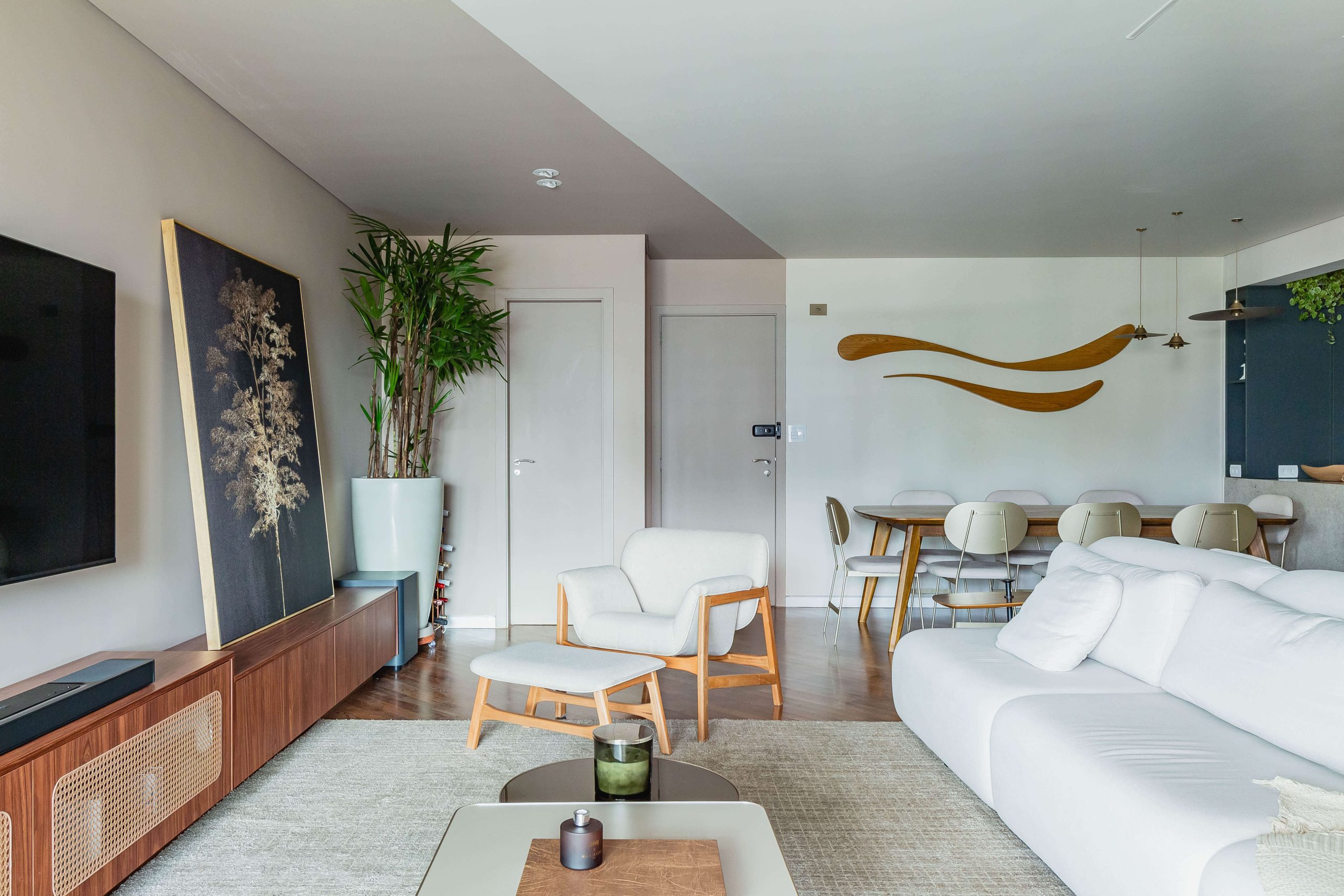 A foto mostra um living amplo com visão para a sala de jantar integrada, destacando o ambiente personalizado com mobiliário em tom escuro, com detalhes nas frentes em porta palha. A televisão fica suspensa na parede. A decoração tem um toque afetivo, com uso de quadros e elementos do decor alusivos a tendência casa de vó para 2025. decoração com mistura de estilos