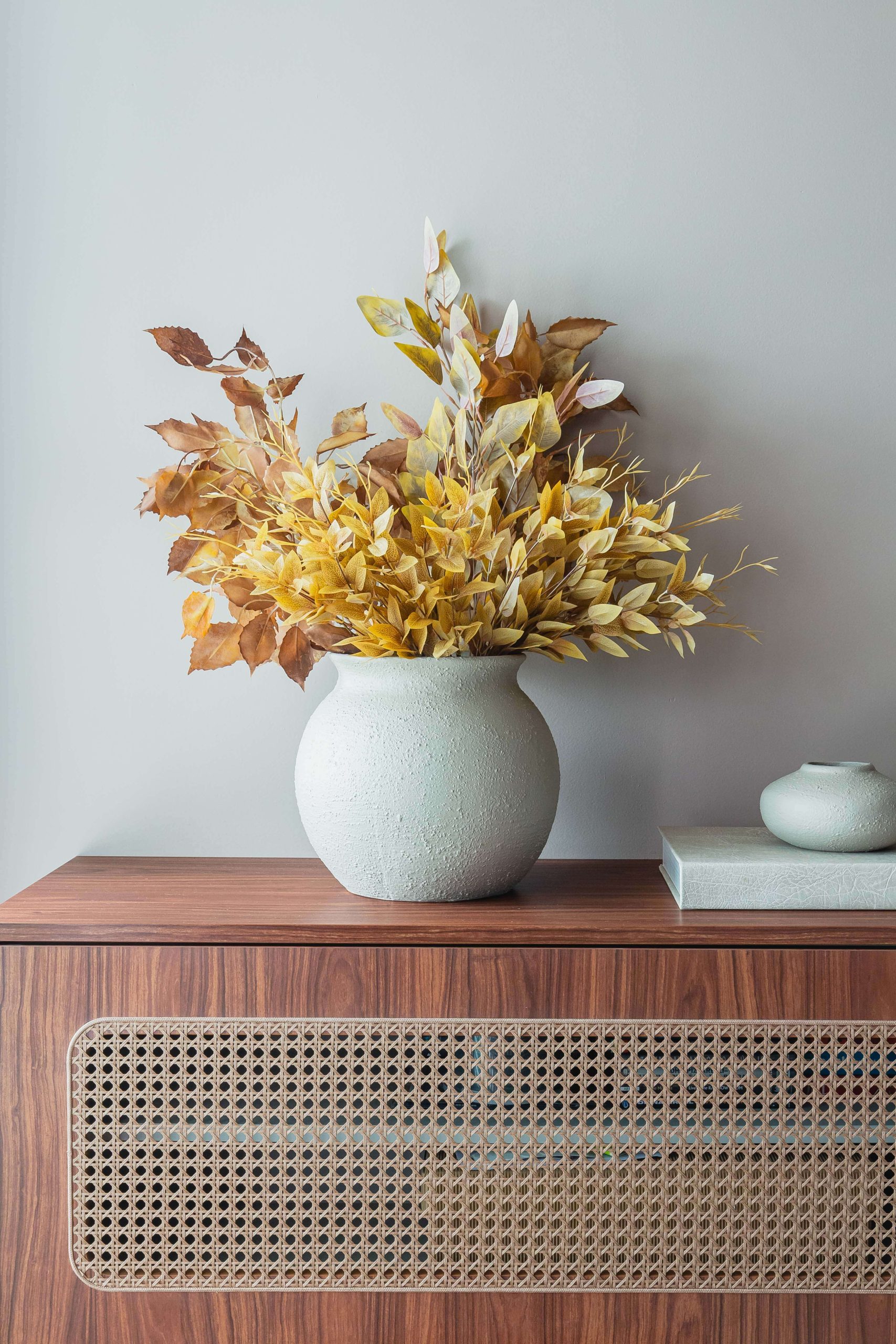 Detalhe do living integrado, com móvel planejado para a sala em tom de madeira escura, com detalhe de porta com palha na cor nogueira. Sobre o móvel está um vaso branco com flores secas em cores outonais. Na lateral um livro branco com um vaso pequeno também branco sobre ele.