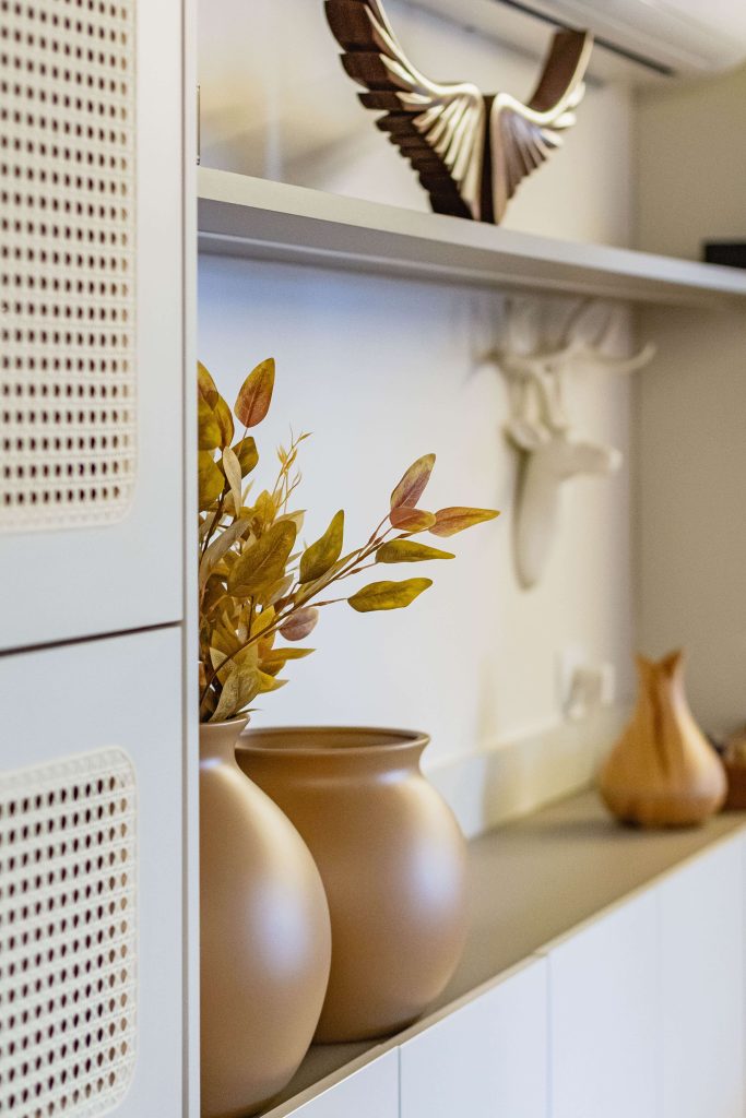 Detalhe de decoração afetiva em um living - sala de estar planejada. Aparece um detalhe das portas com palha indiana, vasos de cerâmica em tons terrosos com folhas secas e esculturas com personalidade e design afetivo.