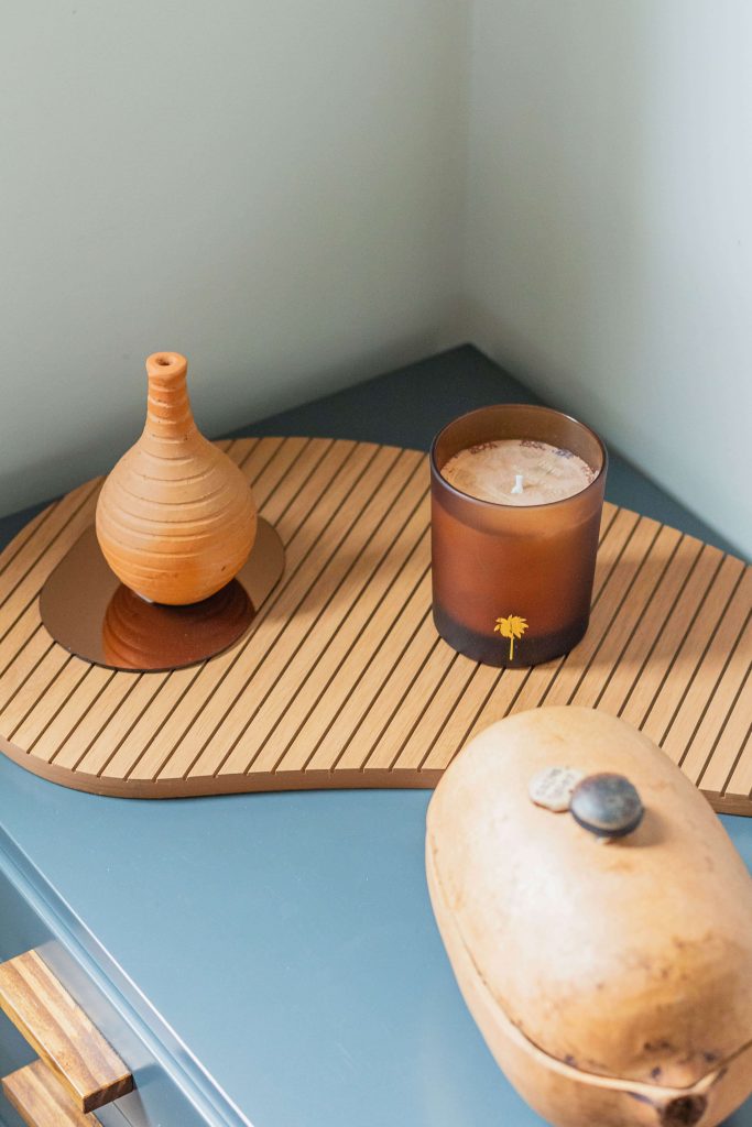 A foto mostra um detalhe da decoração de um quarto planejado, com mobiliário em tom de azul esverdeado. As decorações com formas orgânicas são em tom de madeira e uma vela com vidro âmbar dá um toque de suavidade.