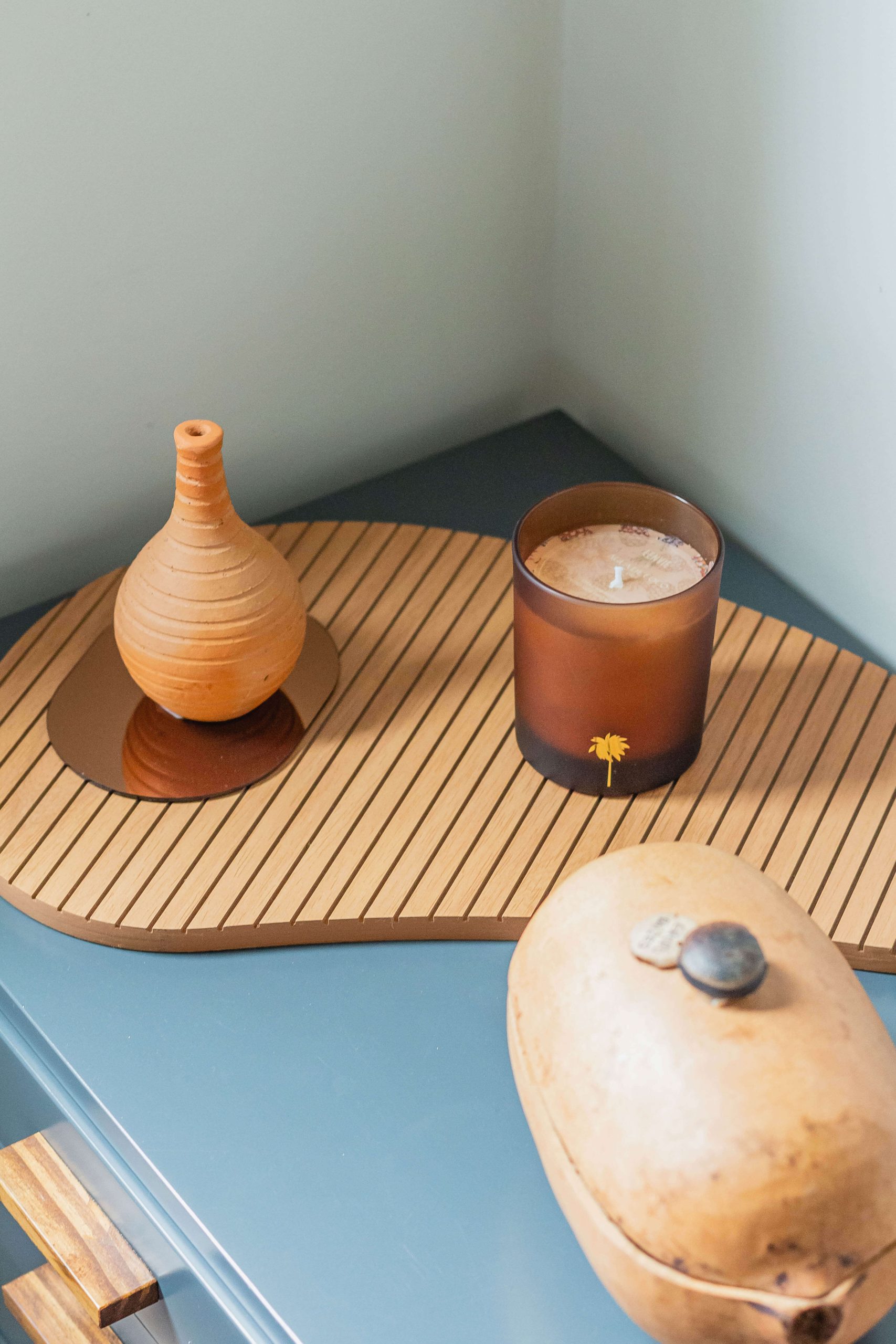 A foto mostra um detalhe da decoração de um quarto planejado, com mobiliário em tom de azul esverdeado. As decorações com formas orgânicas são em tom de madeira e uma vela com vidro âmbar dá um toque de suavidade.