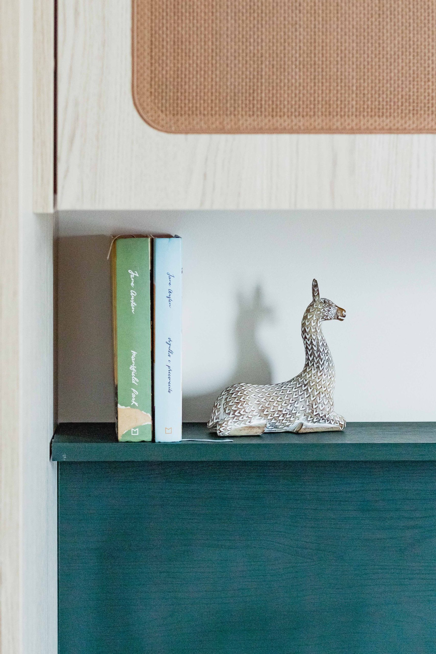 Detalhe de uma prateleira em ambiente de quarto com home office. A foto mostra o móvel planejado em madeira clara linheira, com porta palha de algodão, prateleira em tom de verde profundo com veios de madeira. A decoração é sutil com uma escultura de alce e dois livros.
