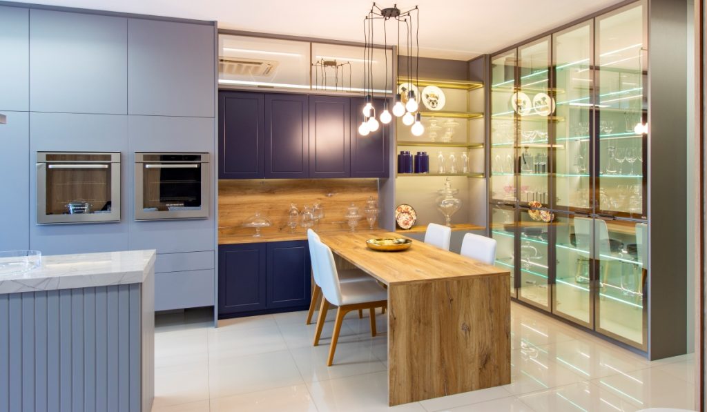 móveis planejados para cozinha com ilha e mesa de jantar em tom de azul e cinza, com detalhes em madeira rústica. Cristaleira com portas de vidro. 