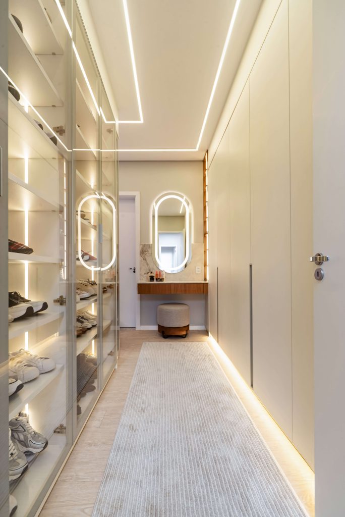 Closet de luxo personalizado, no formato corredor, tendo uma área toda em vidro incolor para colocação da coleção de tênis e calçados do casal. Ao fundo vê-se uma penteadeira com espelho iluminado e a porta que dá acesso ao banheiro da suite master. A marcenaria é em tons claros e o closet é no formato com portas. iluminação embutida