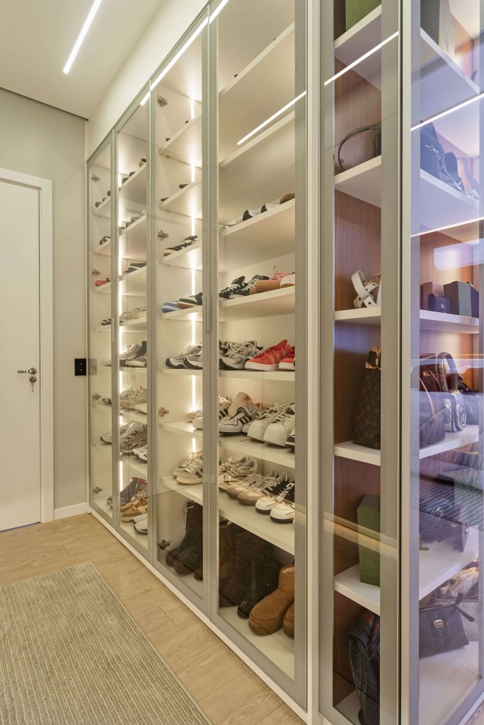 Closet de luxo personalizado, no formato corredor, tendo uma área toda em vidro incolor para colocação da coleção de tênis e calçados do casal. A marcenaria é em tons claros e o closet é no formato com portas. iluminação embutida