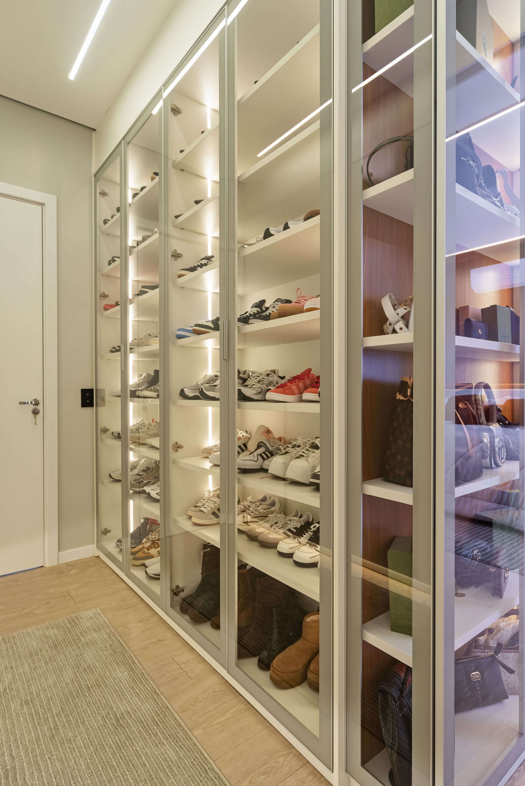 Closet de luxo personalizado, no formato corredor, tendo uma área toda em vidro incolor para colocação da coleção de tênis e calçados do casal. A marcenaria é em tons claros e o closet é no formato com portas.