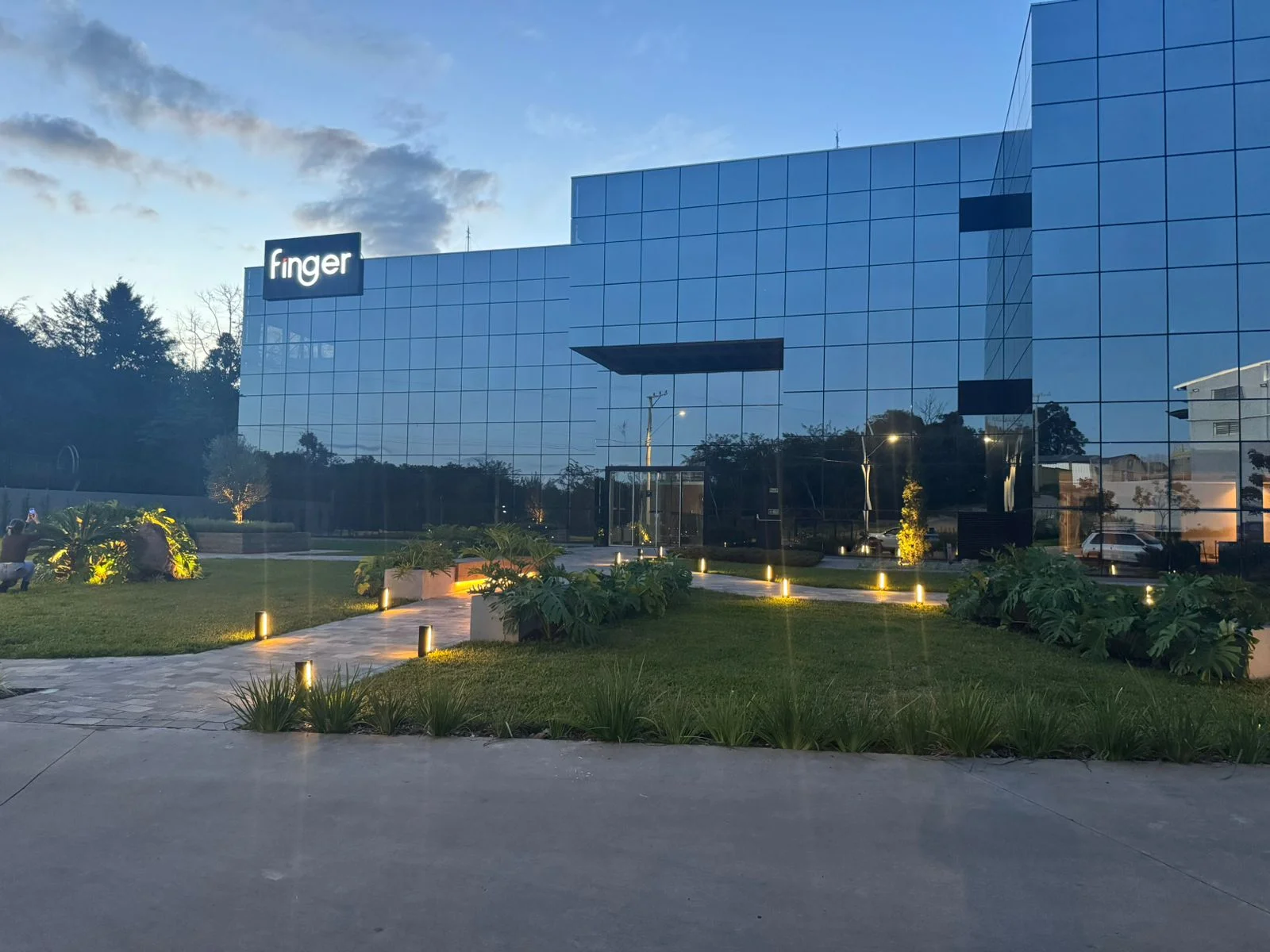 Sede da Finger Móveis Planejados ao entardecer, com arquitetura em vidro espelhado, paisagismo iluminado e logotipo em destaque.