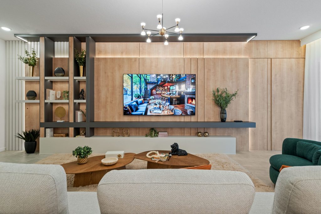 Sala com rack planejado estilo painel suspenso integrado com estante. Ele se destaca por combinar uma bancada flutuante com um grande painel amadeirado em toda a parede, nichos verticais abertos para decoração e iluminação embutida em LED.