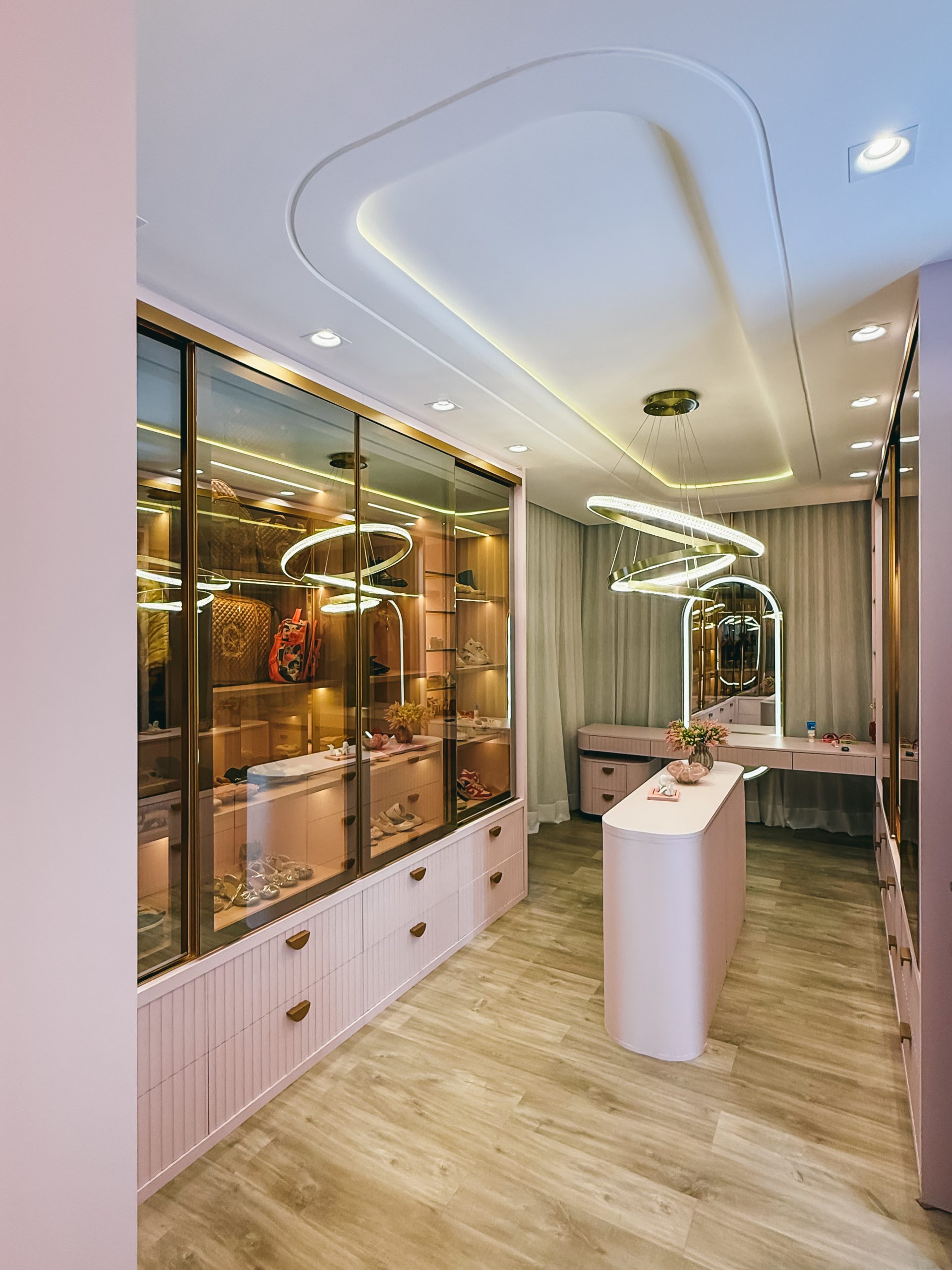 A imagem mostra um closet luxuoso em tons de rosa suave, equipado com armários com portas de vidro transparente e perfis em dourado.
Ao centro, destaca-se uma ilha central oval com cantos arredondados, sob um moderno candelabro lustre com arcos luminosos interligados. Ao fundo, o espaço inclui uma penteadeira com espelho iluminado em arco e cortinas fluidas, sobre um piso em madeira clara.